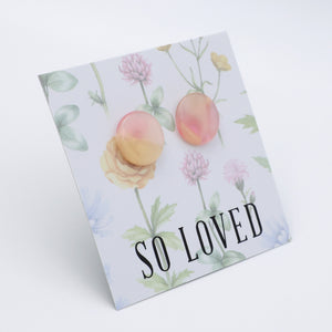 A square printed card or sign with a floral watercolor design, featuring two peach-colored circle earrings pinned on top and the text 'SO LOVED' in large black letters near the bottom. The card is photographed on a clean white background at a slight angle.