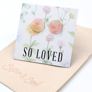 A pair of round, translucent pink and yellow stud earrings displayed on a floral backing card that reads 'SO LOVED', resting on a beige fabric pouch branded 'Sister & Soul'.