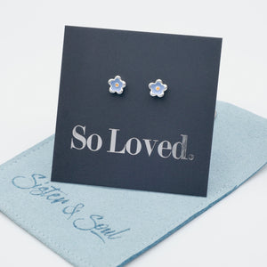 Blue flower earrings with blue and orange enamel detail on a grey card with silver 'So Loved' writing sitting on a blue pouch with 'Sister & Soul' writing.