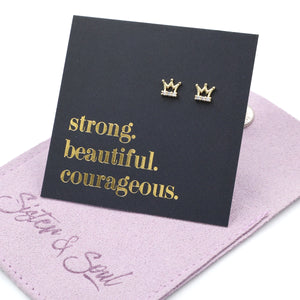 Gold crown earrings on a black card with gold text on a pink surface
