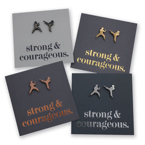 Four square display cards showing small metal stud earrings shaped like martial artists in different poses, each mounted on a dark card with the phrase "strong & courageous." in metallic lettering.