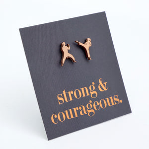A pair of small wooden stud earrings shaped like two martial artists, displayed on a dark gray textured card that reads 'strong & courageous.' in copper-colored lettering, set against a plain white background.