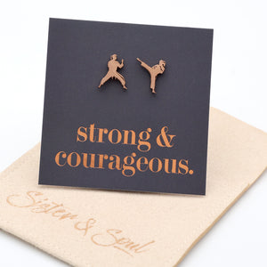 A pair of rose gold metal stud earrings shaped like two martial artists, displayed on a dark charcoal card that reads 'strong & courageous.' with a beige gift pouch underneath on a white background.