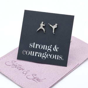 A pair of silver-tone metal stud earrings shaped like two martial artists, displayed on a black square backing card that reads 'strong & courageous.' The card rests on a purple fabric pouch with the words 'Sister & Soul' in script.
