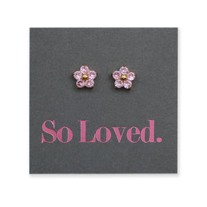 A pair of flower-shaped stud earrings with pink faceted stones and gold centers, displayed on a dark gray card printed with the pink text 'So Loved.'.