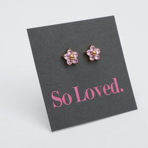 A pair of flower-shaped stud earrings with pink rhinestones and gold centers, displayed on a dark gray card that reads 'So Loved.' in pink text, photographed on a light gray background.