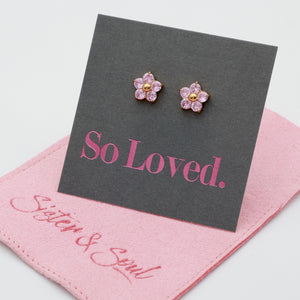 A pair of small flower-shaped stud earrings with pink stones and gold centers, displayed on a dark gray card that reads 'So Loved.' A pink fabric pouch with 'Sister & Soul' branding lies underneath on a white background.