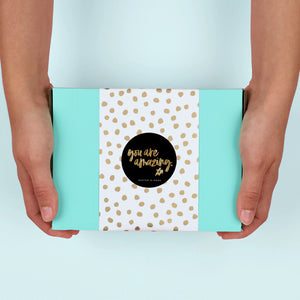 Person holding a turquoise box with a gold polka dot label and 'you are amazing' text.