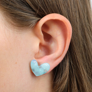 A close-up of a person's ear wearing a small light blue marbled heart-shaped stud earring.
