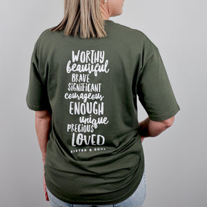 A woman shown from the back wearing an olive green t-shirt with white text on the back that reads 'WORTHY beautiful BRAVE SIGNIFICANT courageous ENOUGH unique precious LOVED' and 'SISTER & SOUL' underneath, against a plain gray background.