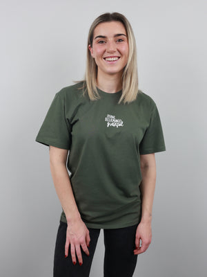 A smiling young woman with blonde hair wearing an olive green t-shirt with a white graphic text print on the left chest, paired with black pants, standing against a solid gray studio background.