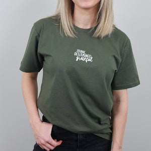 A woman wearing an olive green short-sleeve t-shirt with a white graphic text print on the left chest that reads 'STRONG DETERMINED grateful'.