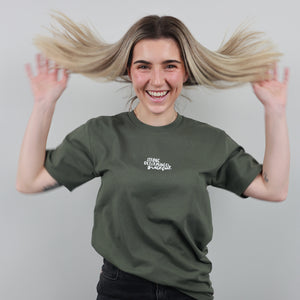 A smiling woman with blonde hair flying out of her hair wears an olive green t-shirt with white text reading 'STRONG BLOOMED STUDIOS' on the left chest, posing against a solid gray background.