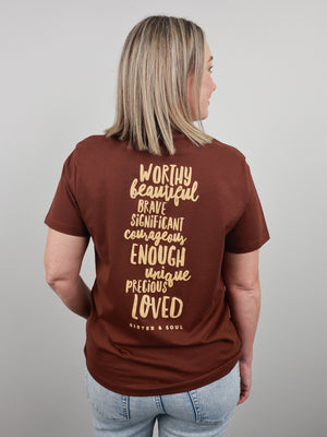 A woman shown from the back wearing a brown short-sleeve graphic T-shirt with light blue jeans, standing against a plain light gray background.
