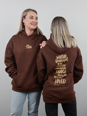 Two women modeling a brown hoodie against a light gray studio background. One shows the front of the hoodie with a small gold chest print, and the other shows the back with a large gold inspirational quote.