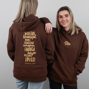 Two women modeling a brown hoodie against a light gray studio background. One shows the back of the hoodie with large gold text reading 'Worthy beautiful brave significant courage enough unique precious loved' and 'Sister a Soul' below.