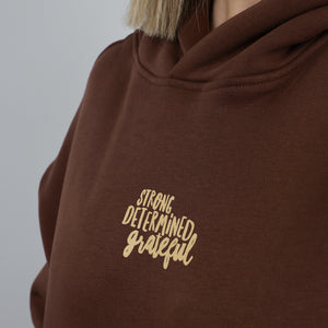 A close-up of a person wearing a dark brown hoodie with the words 'STRONG DETERMINED grateful' printed in a light tan script on the chest, set against a plain light gray background.