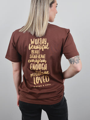 A woman shown from the back wearing a brown short-sleeve t-shirt with a gold typographic quote printed on the back. The text reads 'WORTHY beautiful BRAVE SIGNIFICANT courageous ENOUGH PRECIOUSLY LOVED' and 'SISTER & SOUL'.