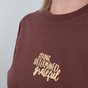A close-up of a person wearing a brown crewneck sweatshirt with the words 'STRONG DETERMINED grateful' printed in a light beige script font on the chest.