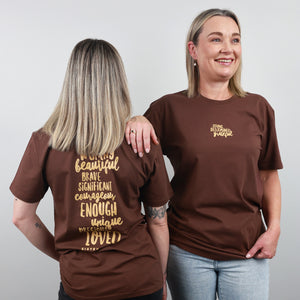 Two women modelling brown graphic T-shirts against a light grey studio background. The woman on the left shows the back of the shirt with gold text reading 'WORLD beautiful BRAVE SIGNIFICANT courageous ENOUGH unique AND CAPABLE LOVED'.