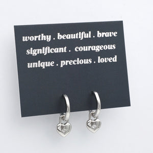 Silver heart-shaped earrings on a black card with positive affirmations on a white background