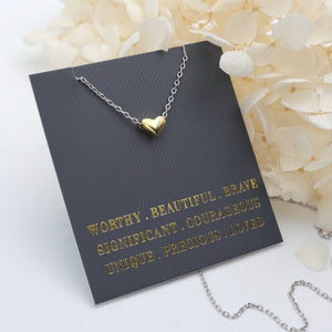 Gold heart necklace on a black card with gold text, placed on a white surface with white flowers.
