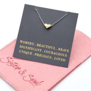 Gold heart necklace on a card with motivational phrases, placed on a pink surface with 'Sister & Swirl' branding.
