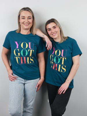 Two women standing side by side wearing matching teal t-shirts with a colorful 'YOU GOT THIS' graphic print, posed against a plain light gray wall.