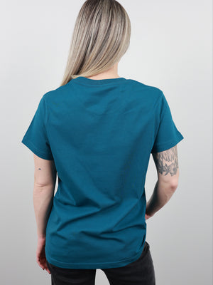 A woman with blonde hair is shown from behind wearing a teal short-sleeve t-shirt and black pants against a light gray background.