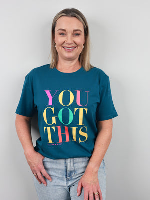 A smiling woman with blonde hair wearing a teal graphic t-shirt that reads 'YOU GOT THIS' in colorful letters, paired with light-wash jeans, standing against a plain light gray background.