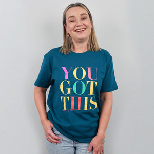 A woman smiling and wearing a teal graphic t-shirt with the text 'YOU GOT THIS' in colorful letters, paired with light-wash jeans, against a plain light gray background.