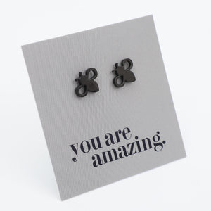 A pair of small black bee-shaped stud earrings displayed on a textured gray square card that reads 'you are amazing.' in black script, photographed on a clean white background.