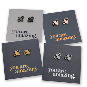 Four pairs of small bee-shaped stud earrings displayed on dark gray cards with the phrase 'you are amazing.' in different metallic colors.