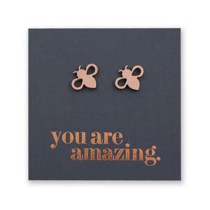 A pair of small bee-shaped stud earrings in a metallic rose gold tone, displayed on a dark charcoal square card with the phrase 'you are amazing.' in matching metallic lettering.