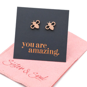 Rose gold bee stud earrings displayed on a black card that reads 'you are amazing.' with a pink fabric pouch underneath on a white background.