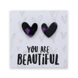 A pair of heart-shaped stud earrings with a glossy black and purple marbled pattern, mounted on a white display card that reads 'YOU ARE BEAUTIFUL' in black text.