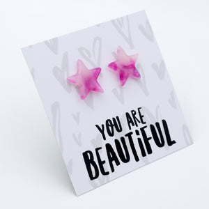 A pair of pink star-shaped stud earrings displayed on a white card that reads 'YOU ARE BEAUTIFUL' in black lettering, photographed on a clean white background.