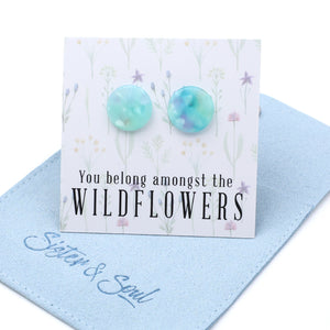 Round stud earrings with a light blue and white marbled pattern, displayed on a white floral backing card that reads 'You belong amongst the WILDFLOWERS'. The card is resting on a light blue suede jewelry pouch embroidered with 'Sister & Soul'.