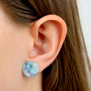 A close-up of a person's ear wearing a round stud earring with a marbled light blue, white, and soft purple pattern.