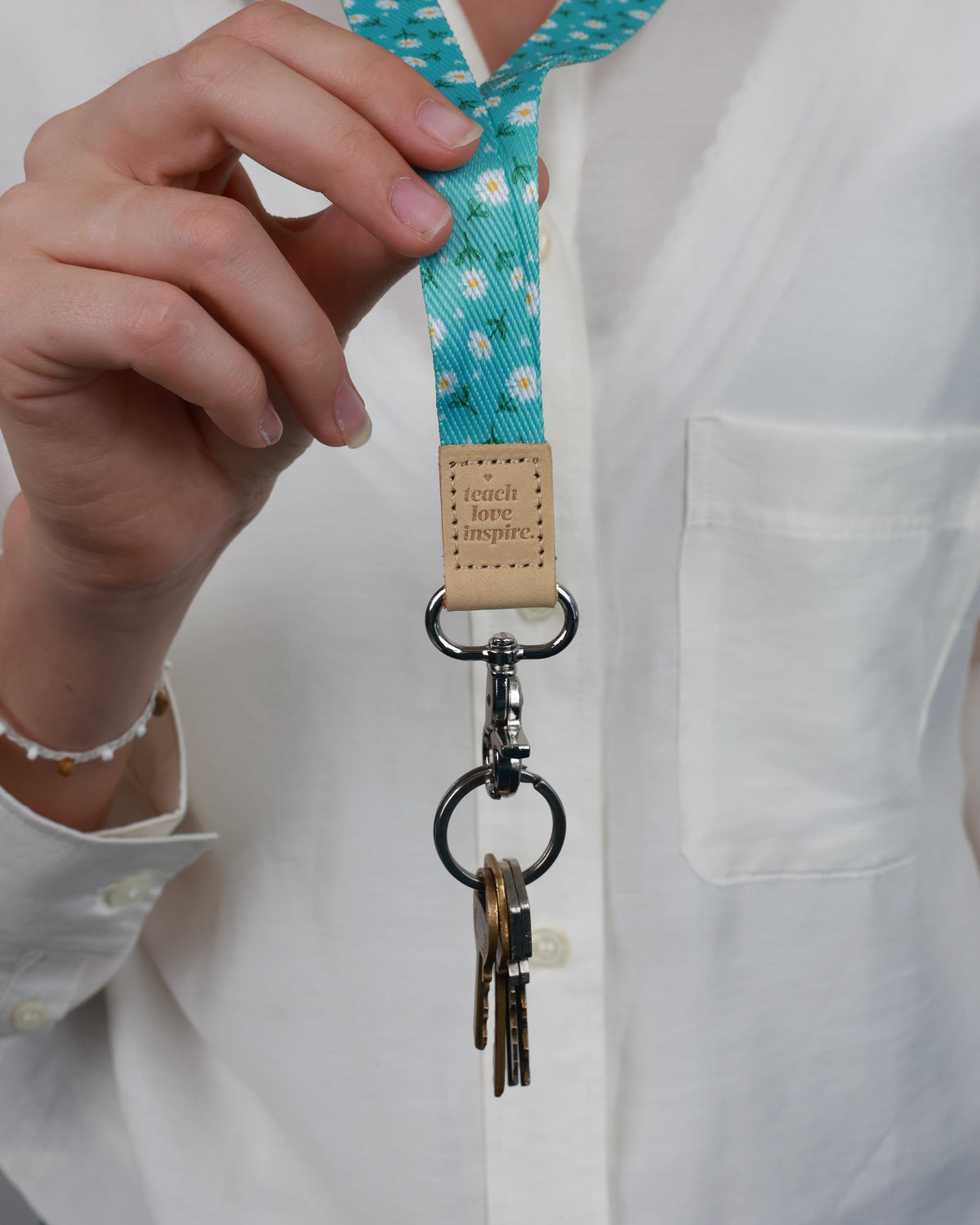 Close-up of the Aqua Daisies lanyard worn over a white shirt, showing its tan Teach Love Inspire tab and silver-tone clasp holding keys.