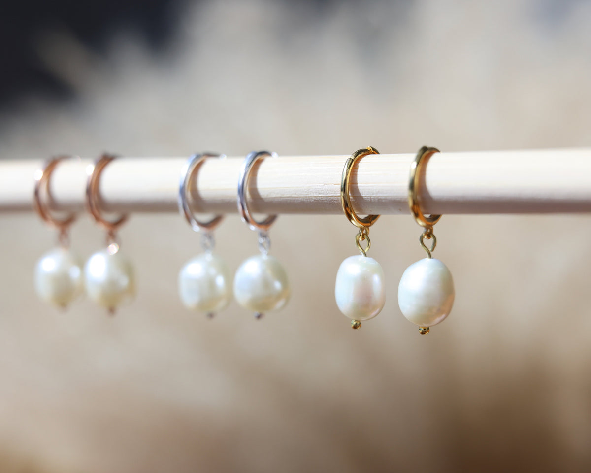 A close-up of several pearl drop earrings hanging from thin metal rings on a white rod, with a soft beige blurred background.