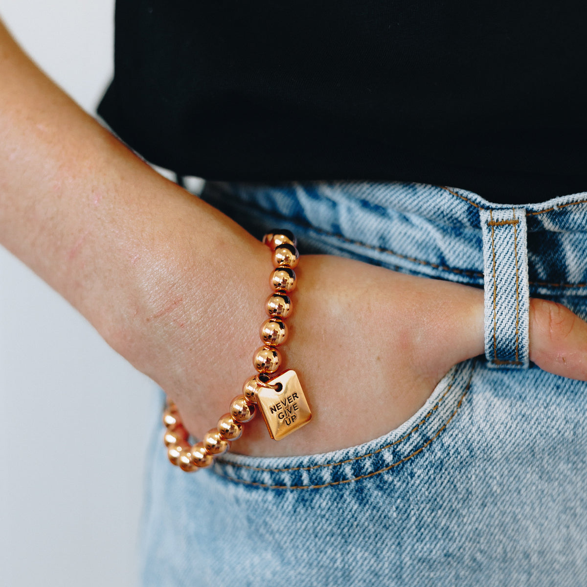 A close-up of a person's hand in a denim pocket wearing a rose gold beaded bracelet with a small engraved tag.