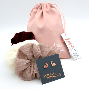 Pink drawstring bag with hair scrunchies, earrings, and a card on a white background