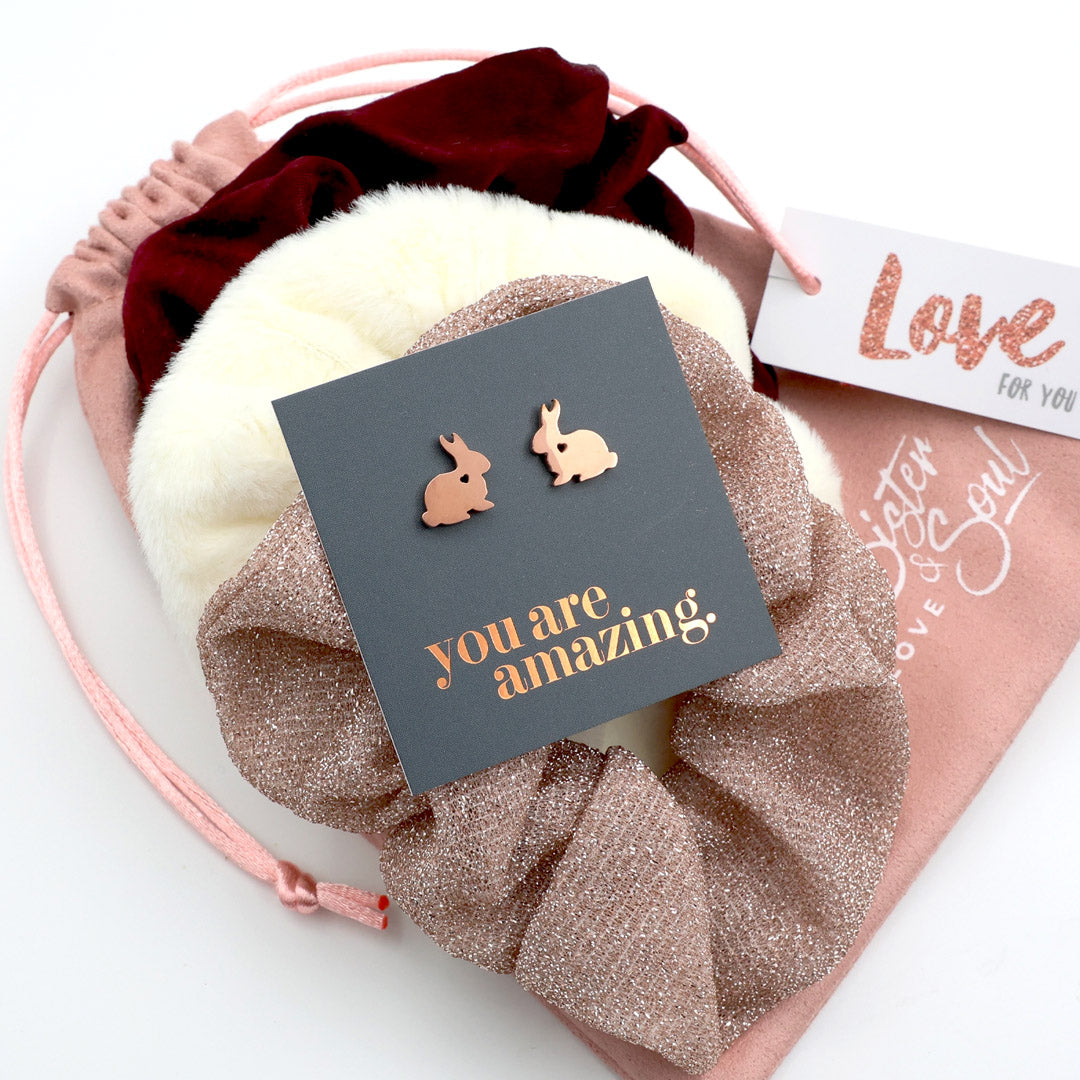 Rose gold bunny earrings on a card with 'you are amazing' text, placed on a decorative pink and white fabric background.