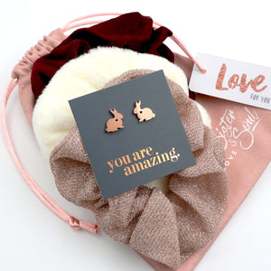 Rose gold bunny earrings on a card with 'you are amazing' text, placed on a decorative pink and white fabric background.