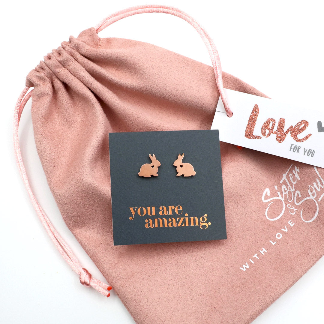 Rose gold bunny earrings on a card with 'you are amazing' text, placed on a pink drawstring bag.