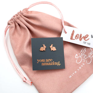 Rose gold bunny earrings on a card with 'you are amazing' text, placed on a pink drawstring bag.