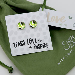 Green and silver earrings on a card with 'Teach Love + Inspire' text, placed on a green fabric background.