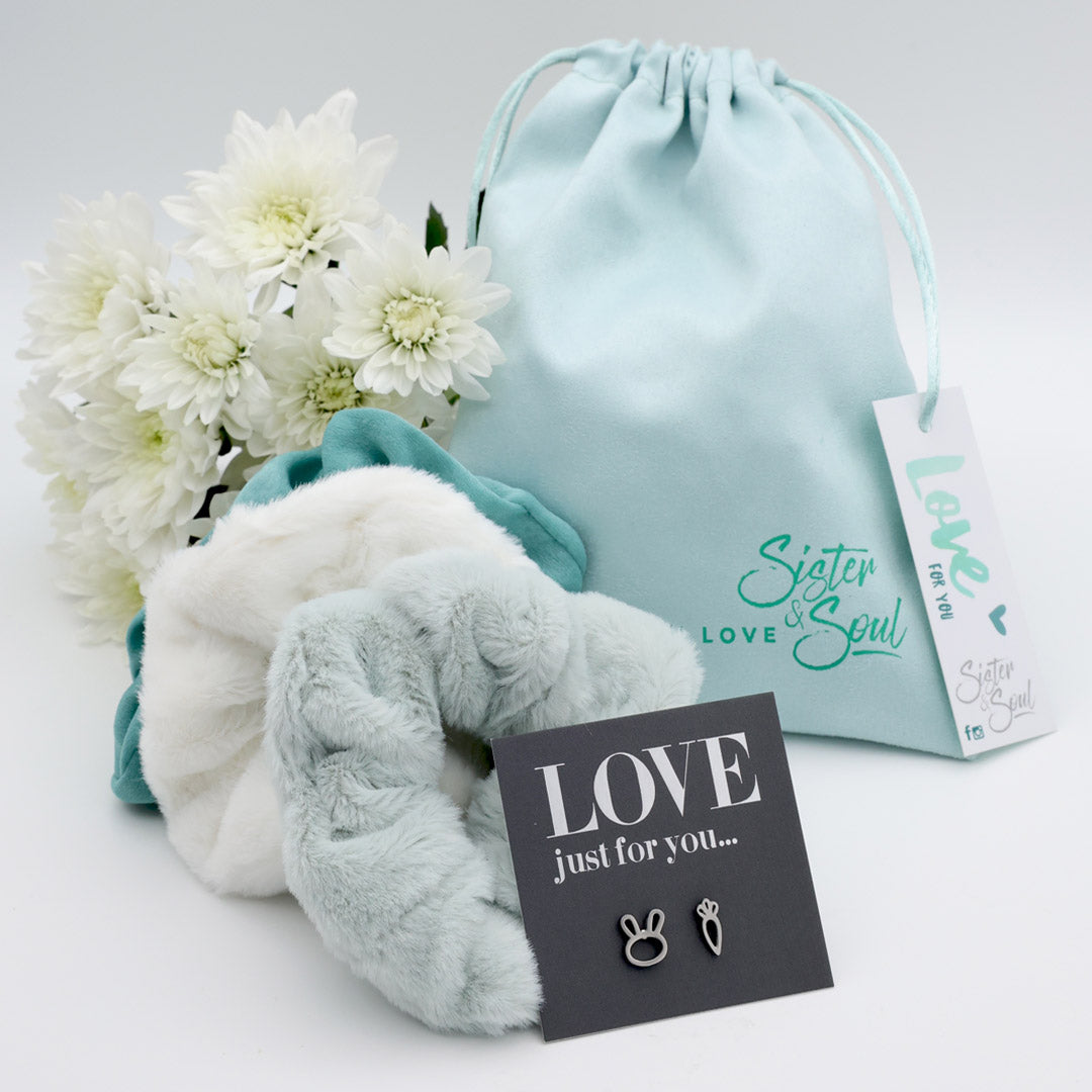 Pair of light blue fuzzy scrunchies with a drawstring bag, flowers, and a card on a white background.