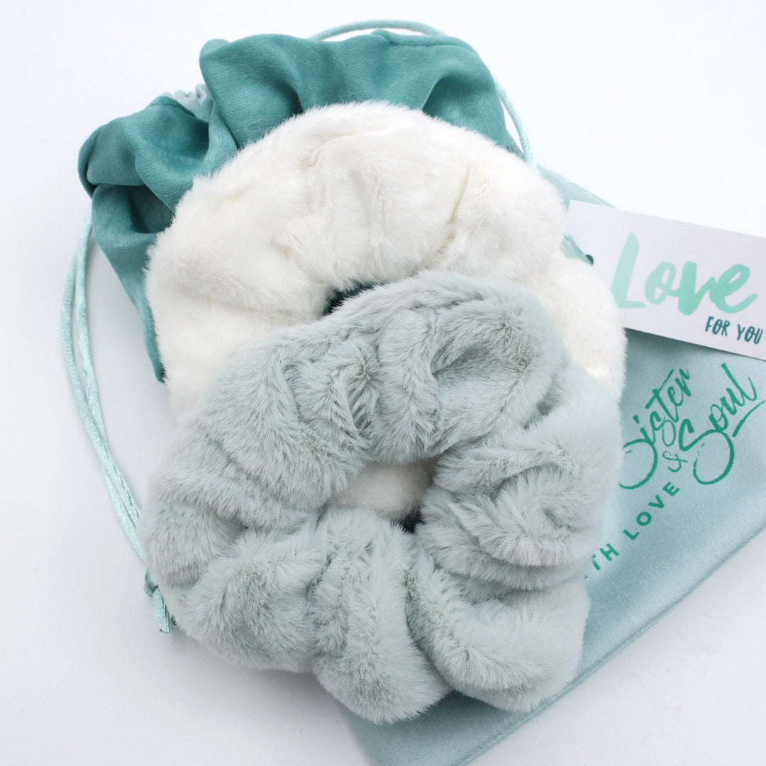 Two plush scrunchies in white and gray on a teal drawstring bag with 'Sister & Soul' branding.